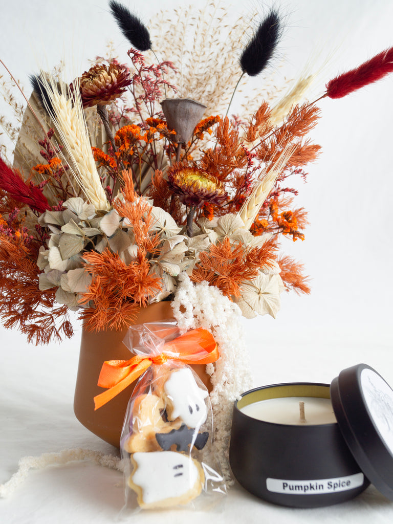 Decorative arrangement with dried flowers, a bag of cookies, and a pumpkin spice candle on a white background